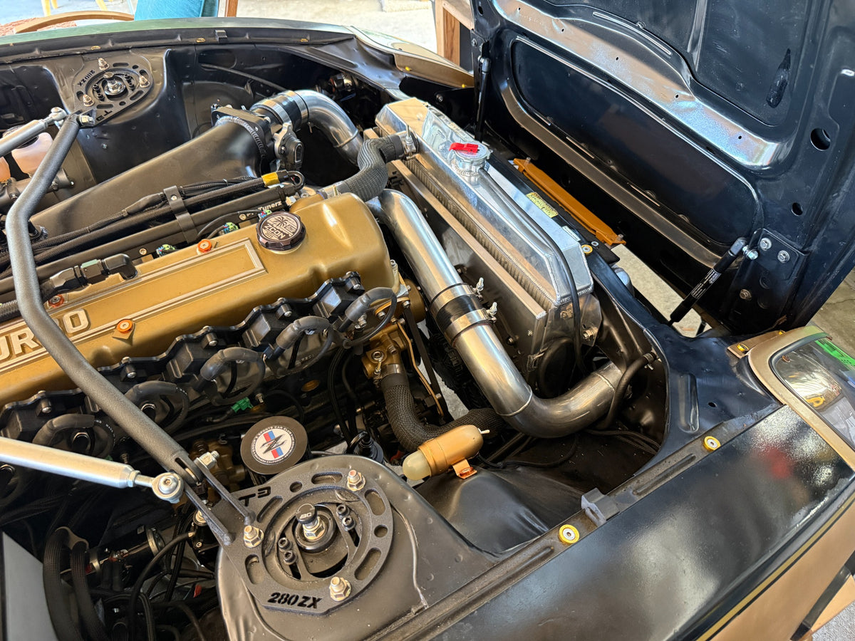Engine bay view of a Datsun 280ZX with the Skillard hood strut kit installed and the hood open.